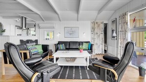 Una sala de estar moderna con techo blanco, sillones de cuero negro, una mesa de centro blanca y un sofá negro con cojines coloridos.