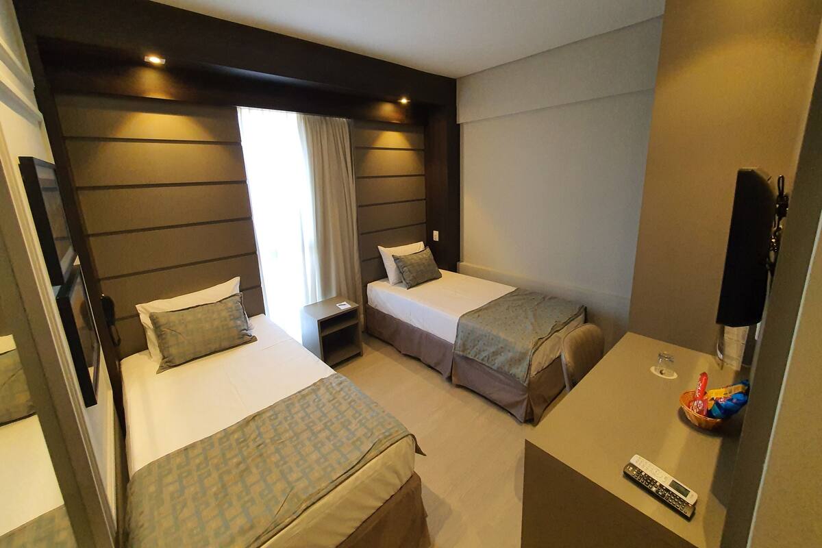 Habitación de hotel con dos camas, una mesita, un televisor y un ventanal con cortinas.