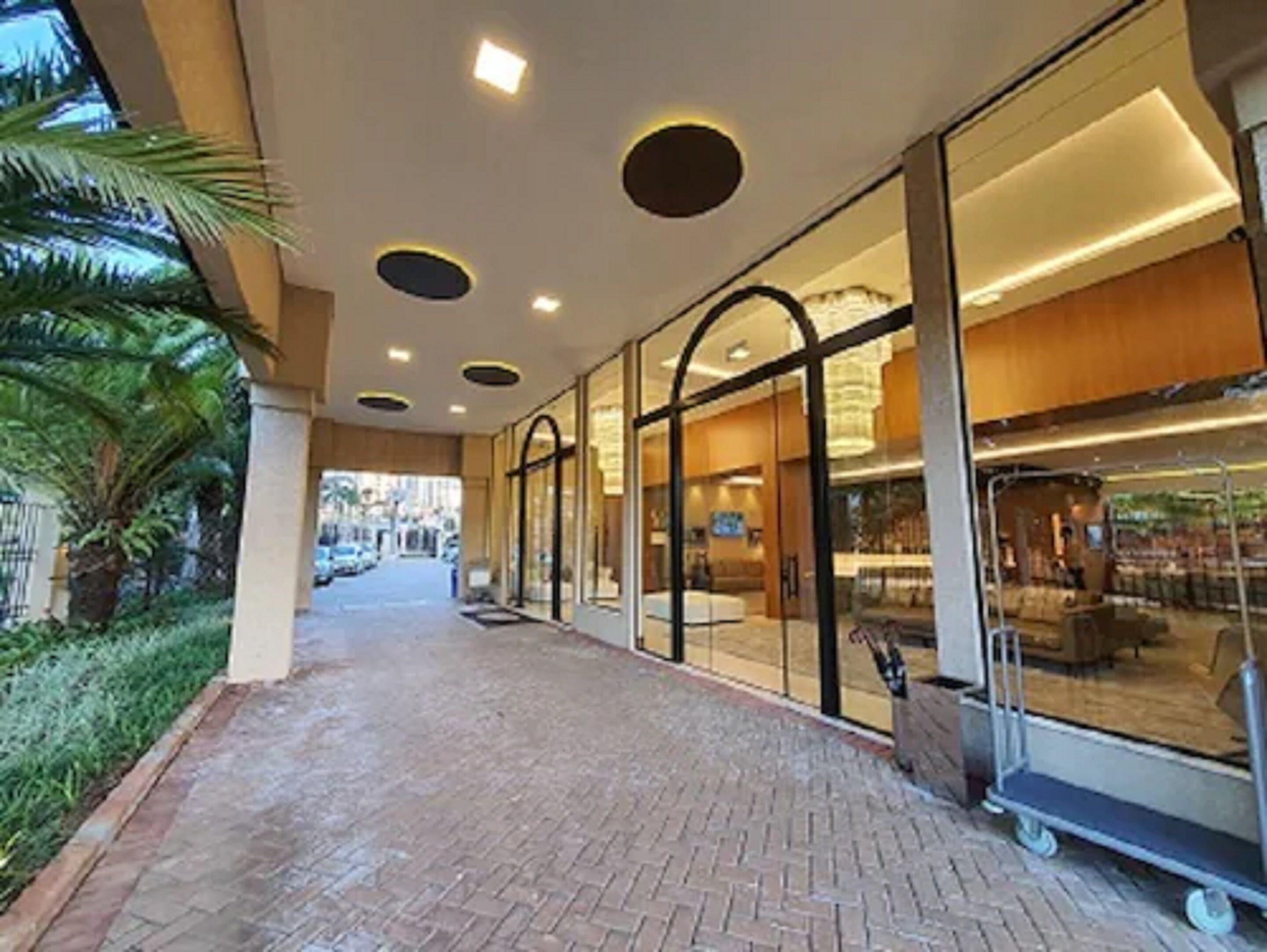 A hotel corridor with a brick floor, glass doors, and a palm tree on the left.