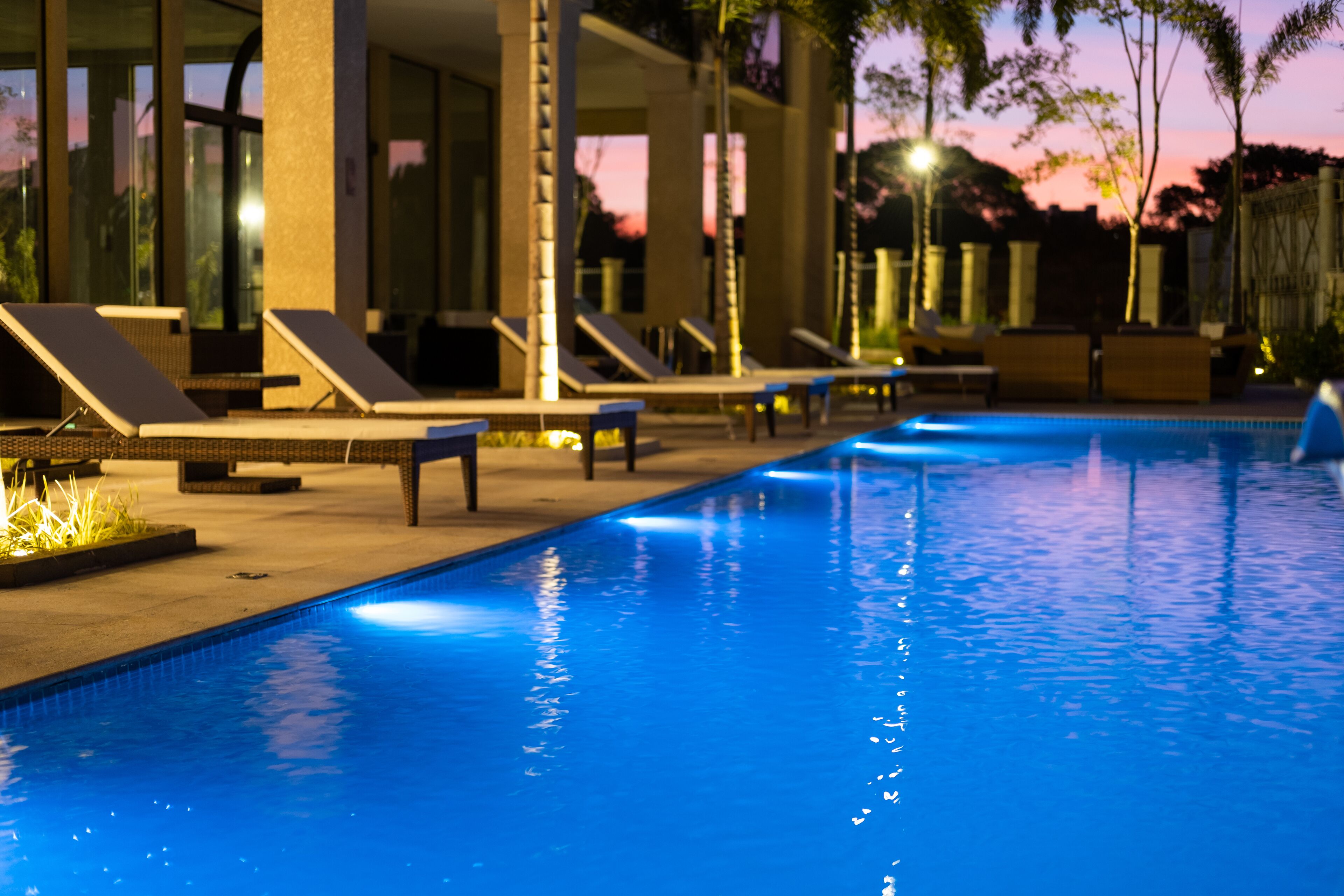 A swimming pool with lounge chairs and a covered area, surrounded by palm trees and a sunset view.