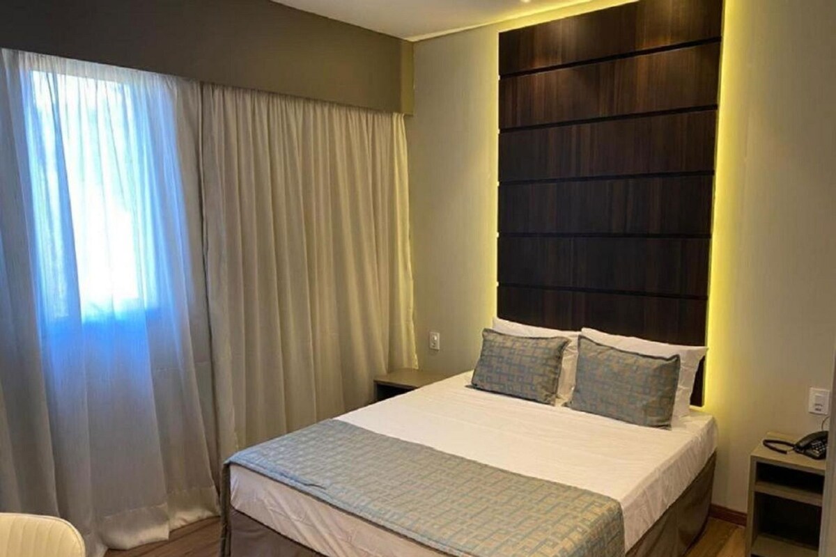 Una habitación de hotel ordenada con una cama grande, una mesita de noche con teléfono y una ventana con cortinas.