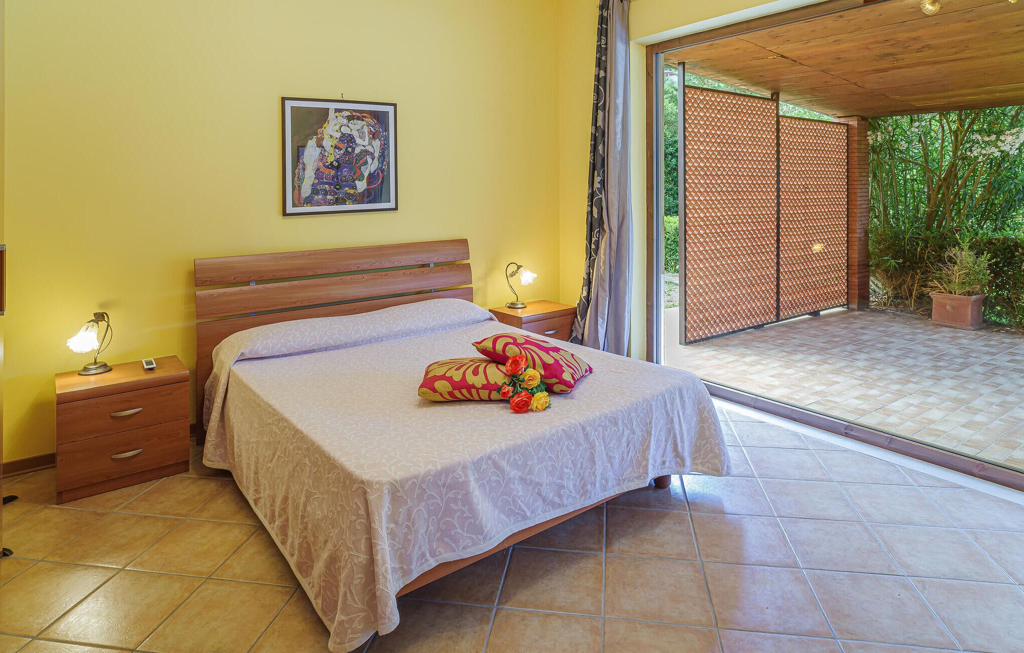 A bedroom with a bed, bedside tables, a lamp, a framed picture on the wall, and a view of an outdoor area with a wooden ceiling.