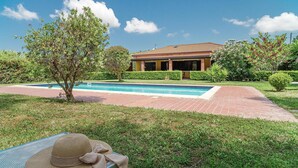 Zona de piscina con una casa al fondo, rodeada de vegetación y un cielo despejado.