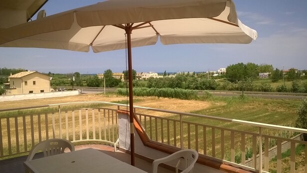 A balcony with a table and chairs, a large umbrella, and a view of a road and buildings in the distance. - Self-catering single room for rent with a private bathroom (Marina di Montenero di Bisaccia)