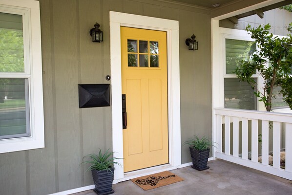A yellow front door with a mailbox, two potted plants, a doormat, and a white porch railing.