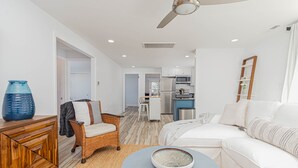 Ein modernes Wohnzimmer mit einem weißen Sofa, einem Korbstuhl, einem Couchtisch mit einer Schale und einer Küche im Hintergrund.
