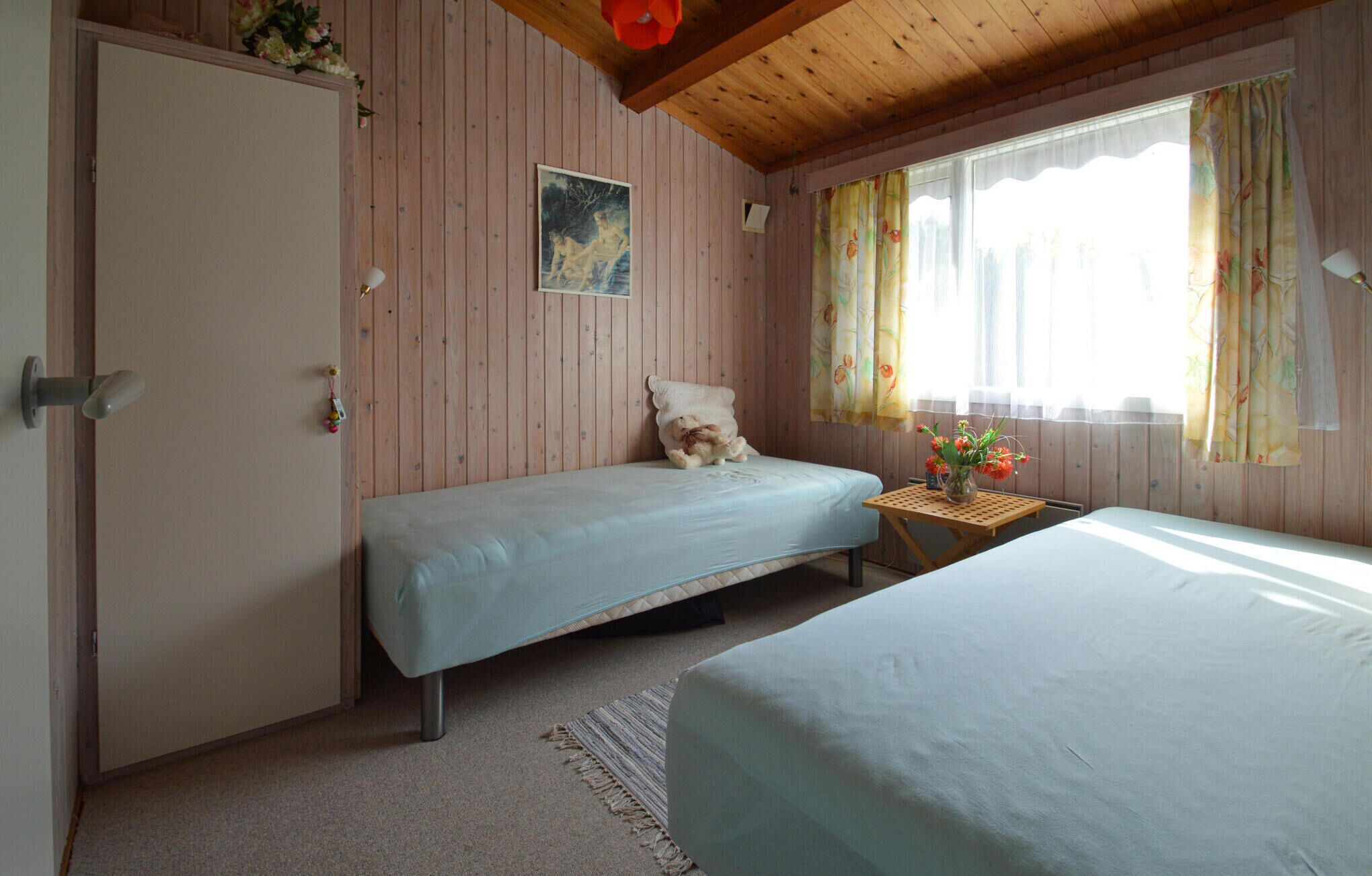 Une chambre avec deux lits simples, des murs en bois, un plafond en bois, une petite table avec un vase et une fenêtre avec des rideaux.
