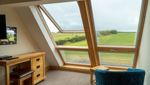 Een kamer met een schuine balkenlaag, een houten bureau met lades, een bankje en een groot raam dat uitzicht biedt op een groen veld.