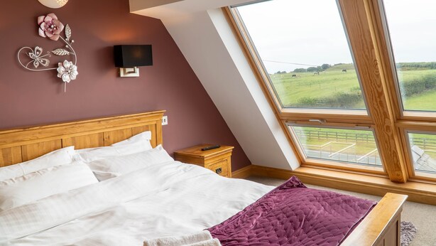 A bedroom with a sloped ceiling, a large bed with white bedding, a purple throw blanket, a wooden nightstand, a wall-mounted TV, and a window with a view of a green field. - Primrose Cottage -  2 Bed cottage with Hot Tub, walk to the beach. (Ulverston)