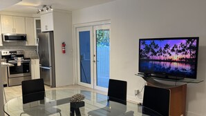 Moderni keittiö, jossa on ruostumattomasta teräksestä valmistetut kodinkoneet, ruokailutila lasipöydällä ja mustilla tuoleilla sekä televisio, jolla näkyy trooppinen auringonlaskukuva.