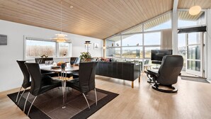 A modern living room with a wooden ceiling, large windows, a black sofa, a dining table with chairs, and a TV.