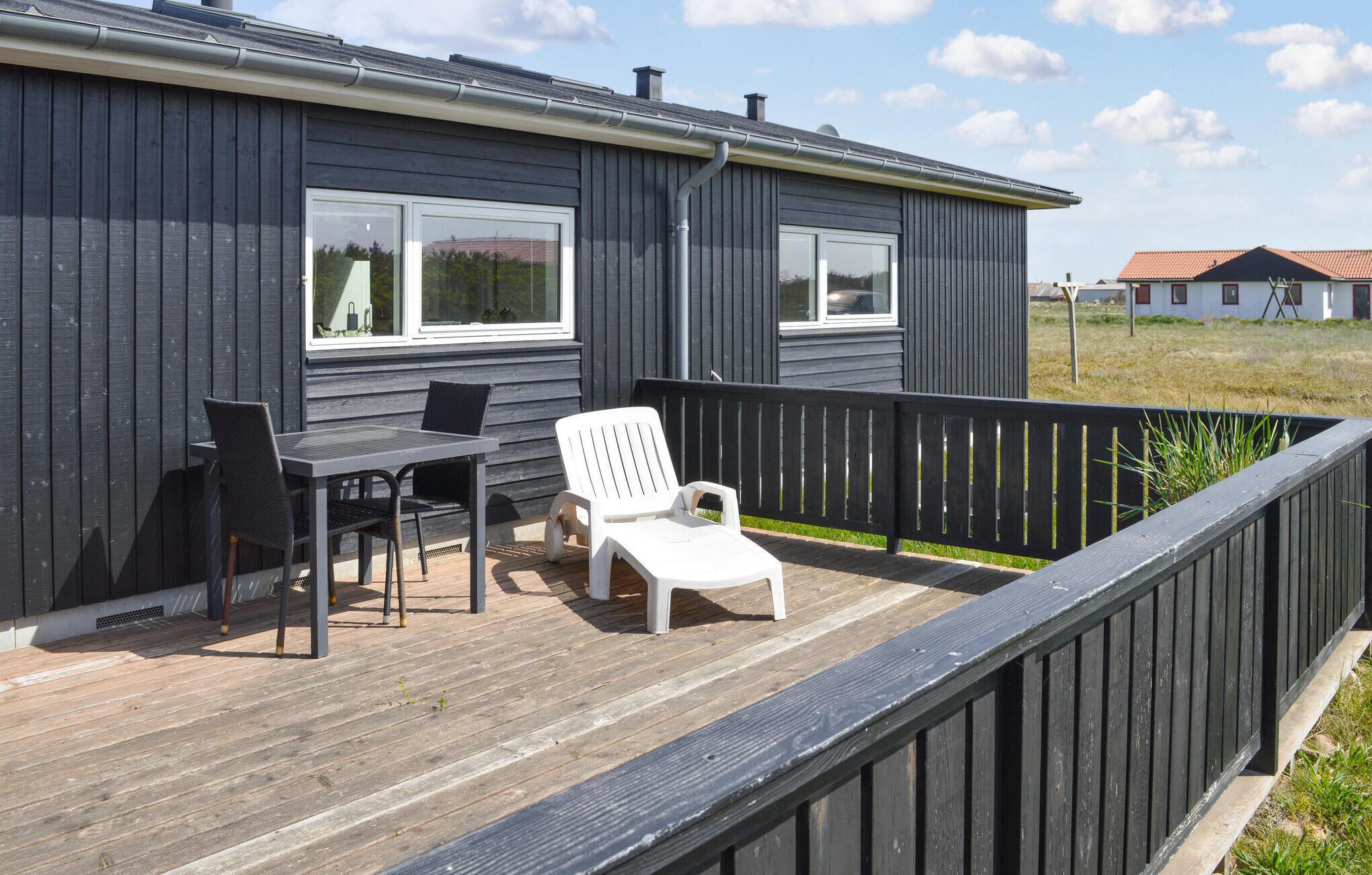 A dark wooden house with a deck featuring a table and chairs, a white lounge chair, and a black railing.