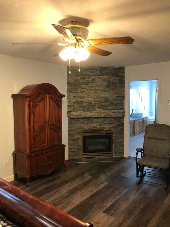 A living room with a stone fireplace, a wooden cabinet, a ceiling fan, and a chair.