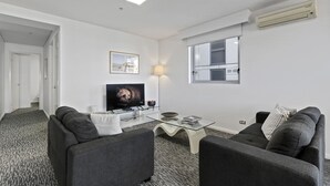 Een moderne woonkamer met een flatscreen televisie, twee grijze banken, een glazen salontafel en een lamp.