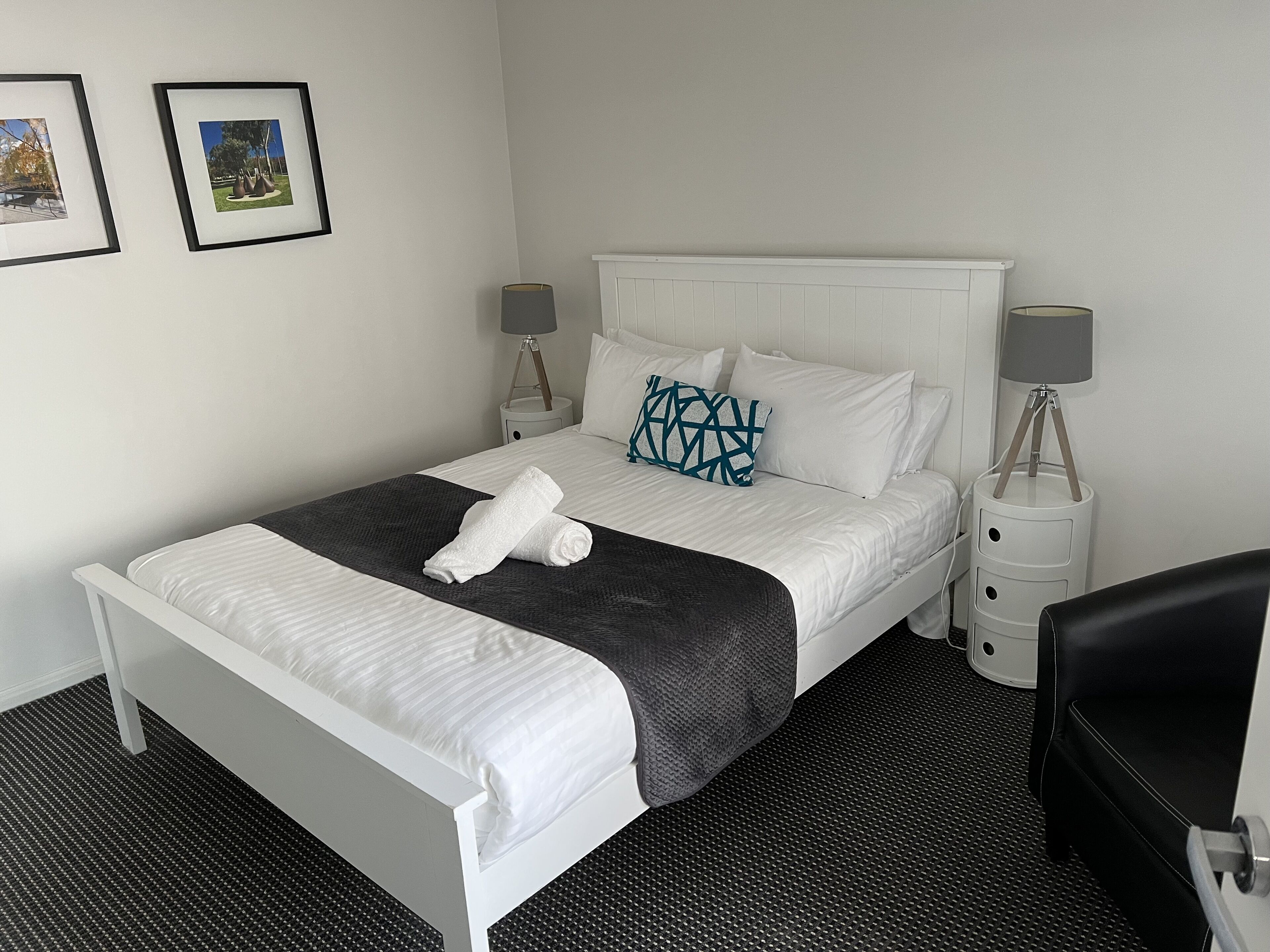 A neatly made bed with a white headboard, a grey blanket, and a rolled white towel. There are two bedside tables with lamps, a black chair, and two framed pictures on the wall.