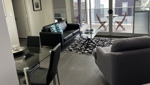 Een moderne woonkamer met een glazen eettafel, een zwarte leren bank en een grijze fauteuil. Er is een groot raam met uitzicht op de stad.