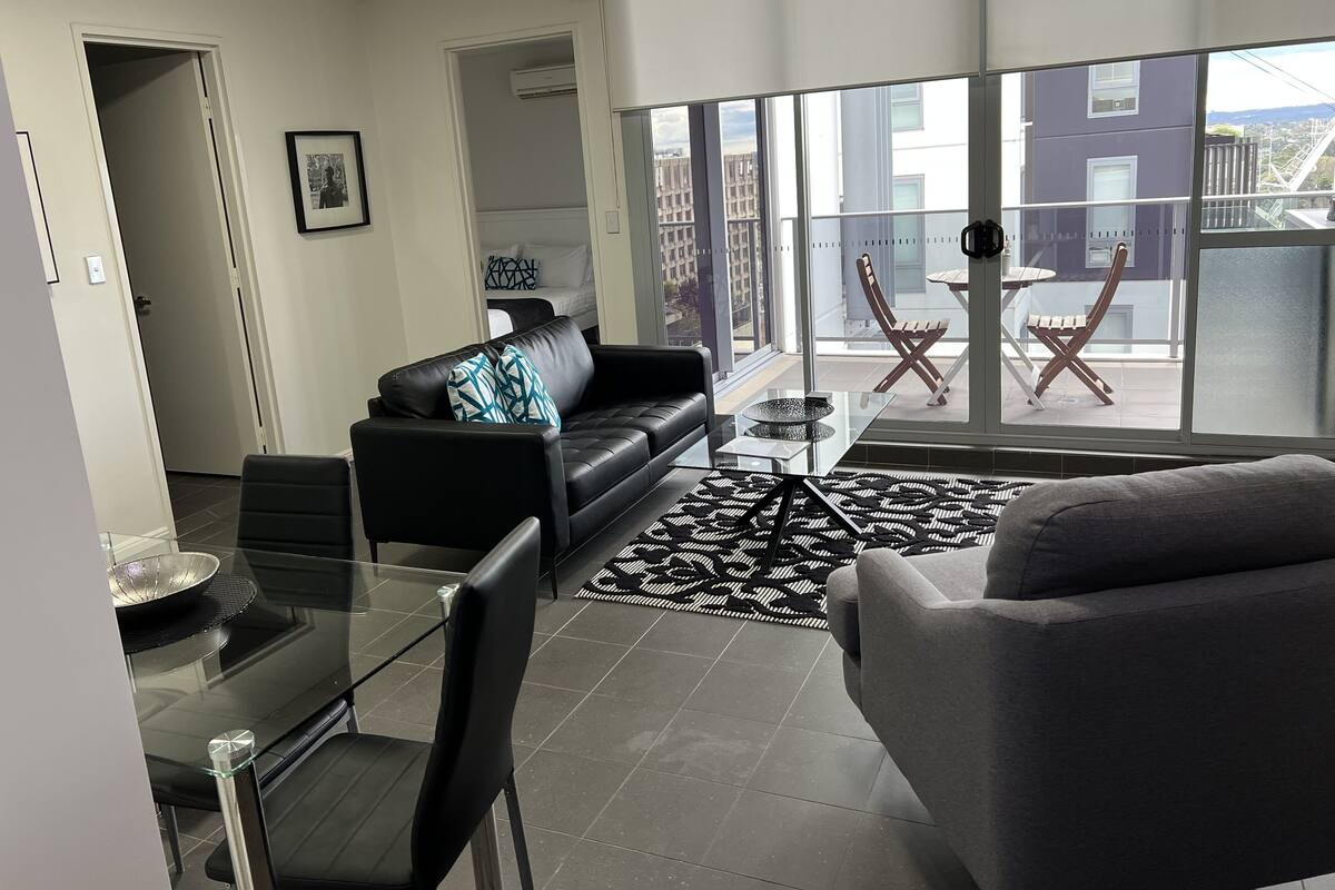 A modern living room with a glass dining table, a black leather sofa, and a grey armchair. There is a large window offering a view of the city.