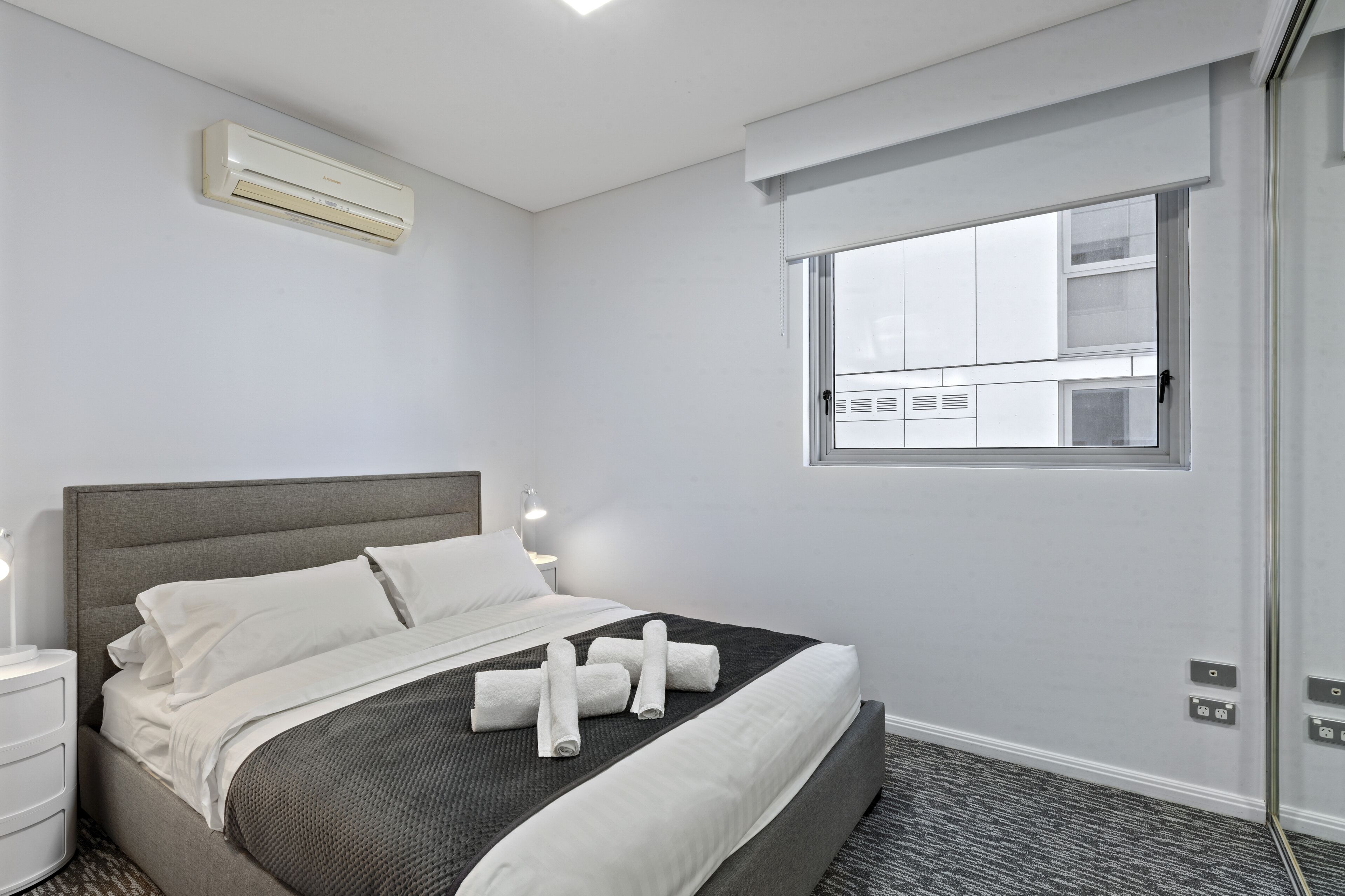 Una habitación de hotel con cama, ventana, aire acondicionado y mesita de noche.