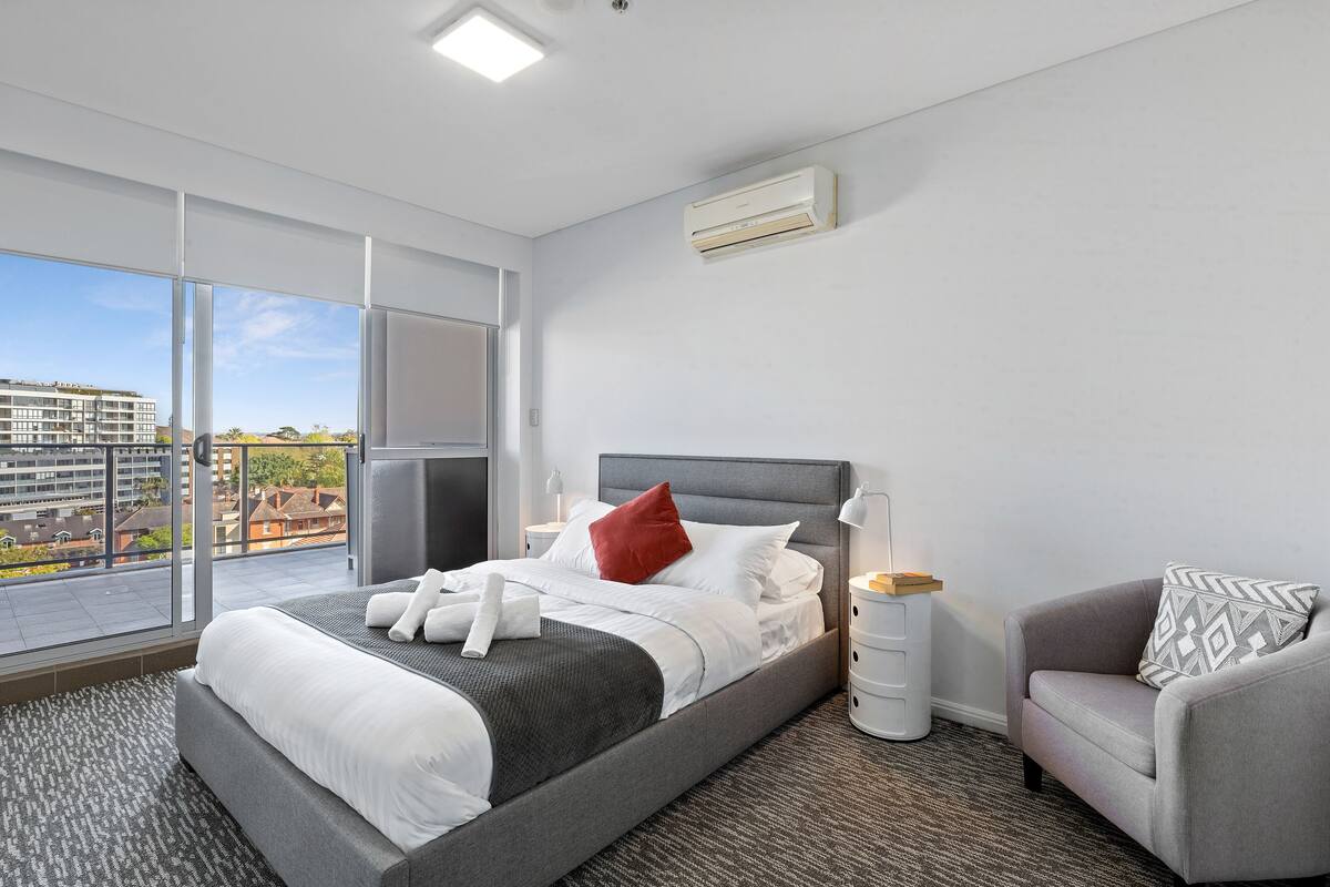 A modern hotel room with a large bed, a grey sofa, and a city view.