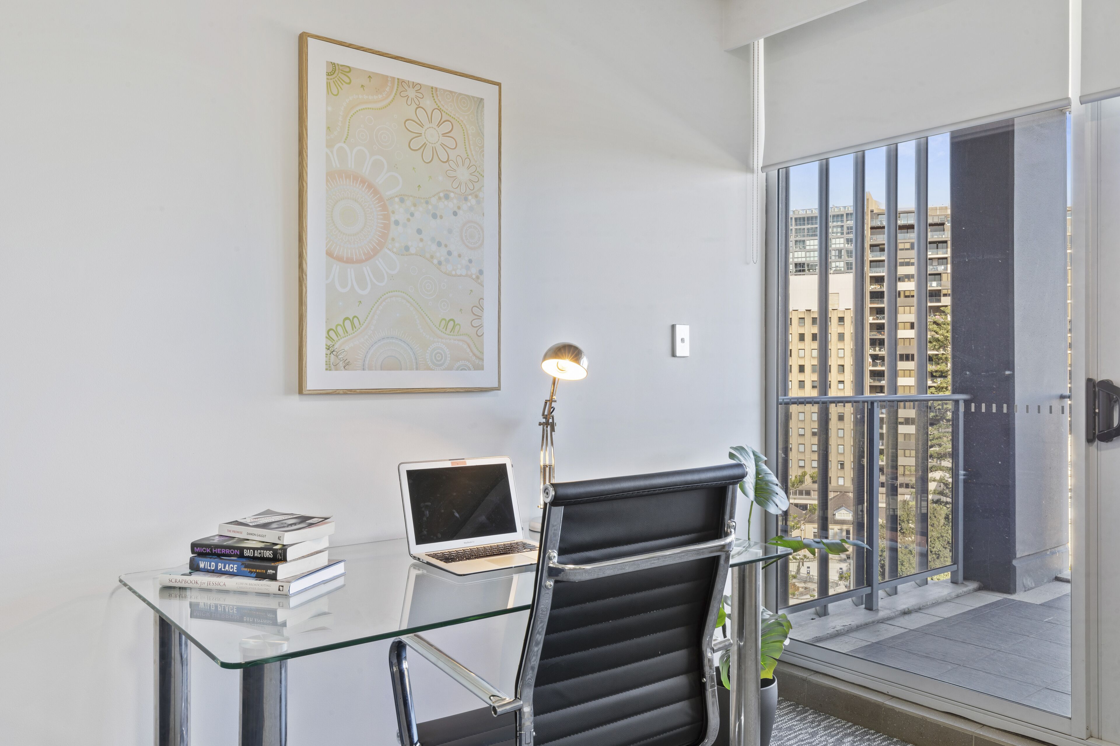 Un espace de bureau moderne avec un bureau en verre, un ordinateur portable, un fauteuil de bureau noir, une lampe et une œuvre d’art encadrée accrochée au mur.