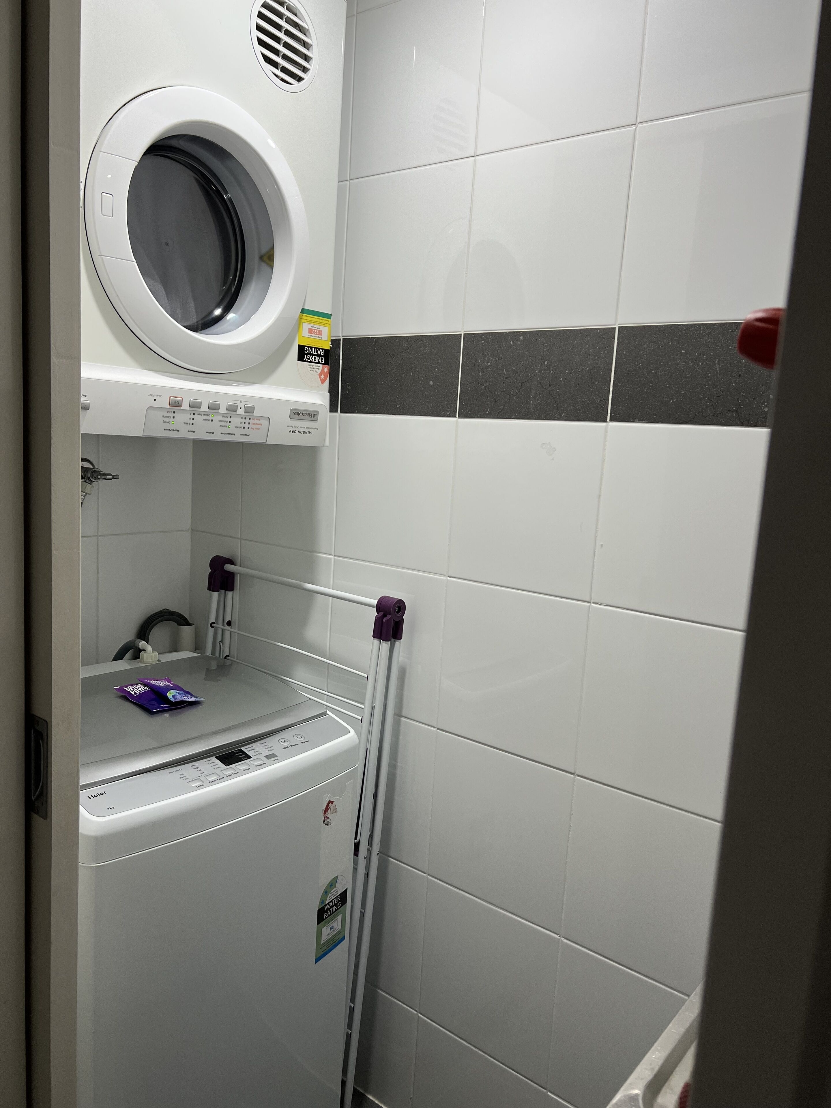 A compact laundry area with a front-loading washer and dryer, a small shelf with a detergent pack, and a towel rack.