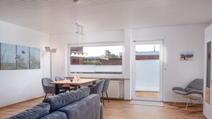 Een moderne woonkamer met een grijze bank, een houten eettafel met stoelen, een radiator en een groot raam met zonwering.