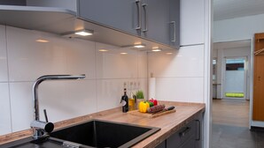 Een moderne keuken met een zwarte spoelbak, een roestvrijstalen kraan en een houten aanrecht. Er zijn kastjes met zilveren handgrepen en zicht op een gang met een houten deur.