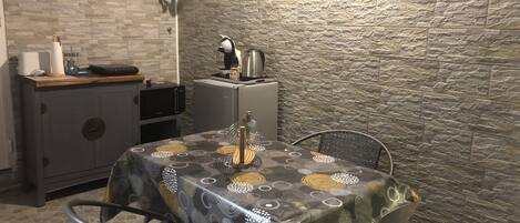 A compact kitchen with a microwave, a coffee maker, and a small refrigerator. There is a table with a patterned tablecloth and two chairs. The kitchen has a stone wall and a wooden ceiling.