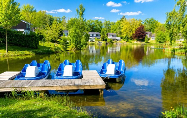 A wooden dock with three blue paddle boats, a calm lake, and houses in the background.