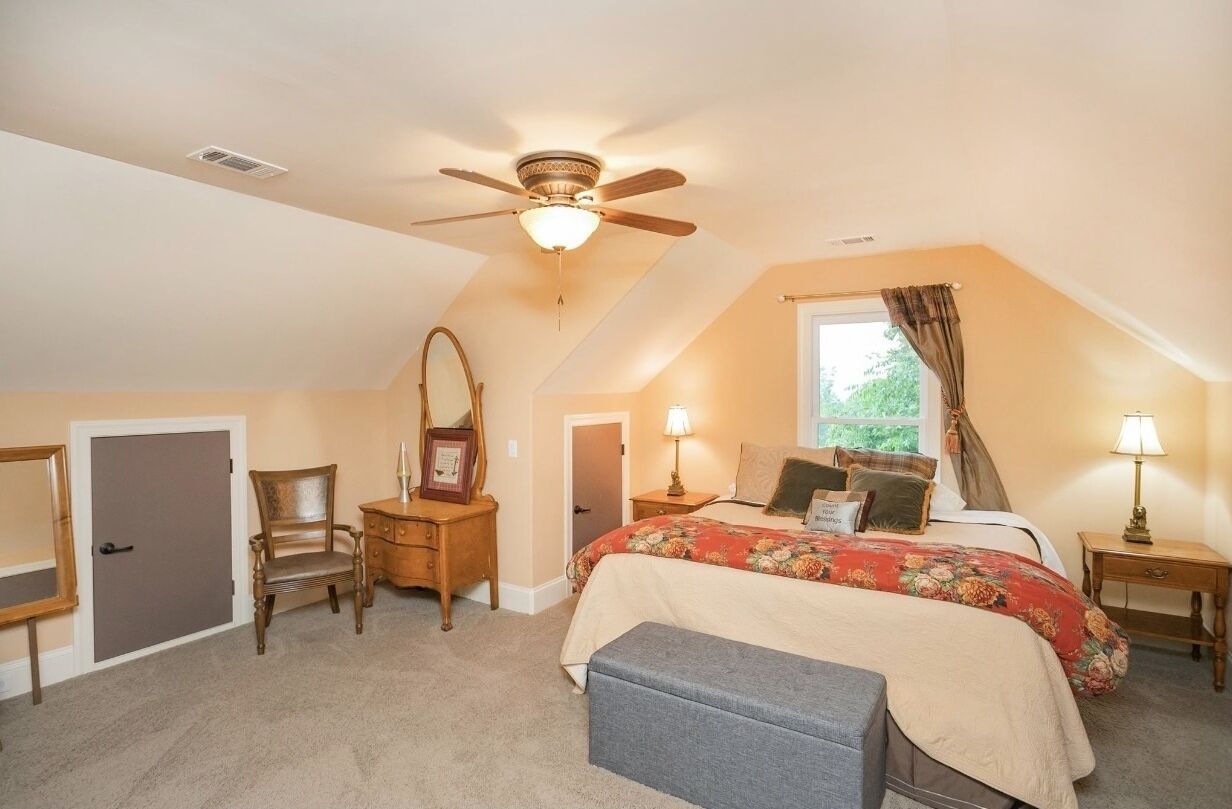 A bedroom with a sloped ceiling, a bed with a floral bedspread, a wooden chair, a nightstand with a lamp, and a ceiling fan.
