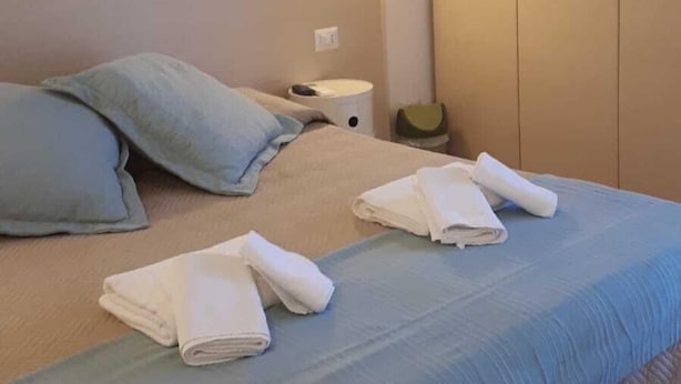 A neatly made bed with pillows, a bedside table with a lamp, and a wardrobe. - Holidayfever (Alghero)