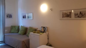 A small, cozy room with a bed, a nightstand, and a refrigerator. There are two framed pictures on the wall and a light fixture above the nightstand. - Holidayfever (Alghero)