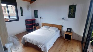A bedroom with a wooden ceiling, a bed with white bedding, a standing fan, a small bedside table, and a window with blinds.