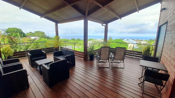 A covered outdoor seating area with wooden flooring and metal chairs, overlooking a lush landscape and the sea.