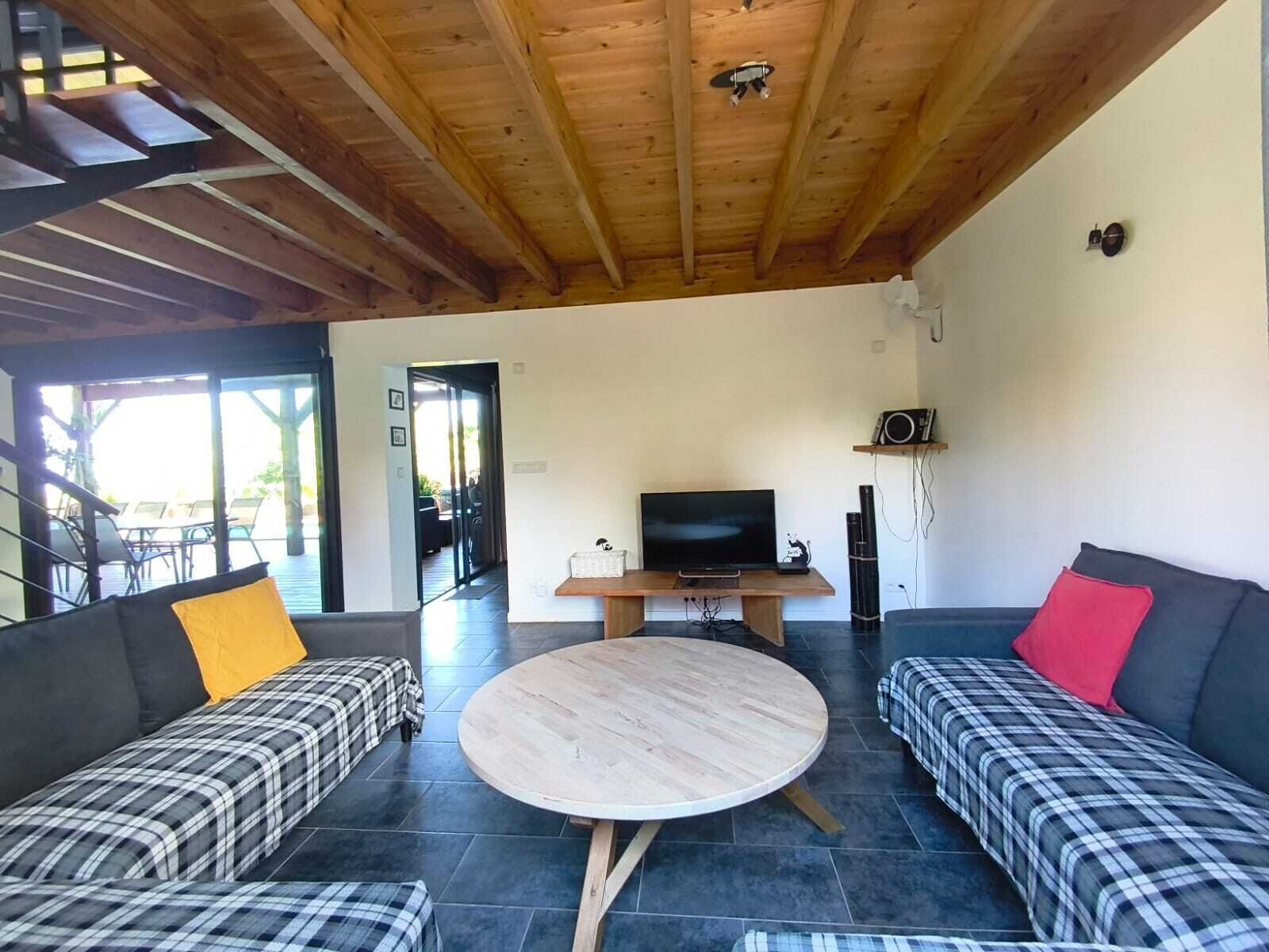 A modern living room with a wooden ceiling, a round coffee table, two sofas with plaid bedding, a flat-screen TV, and a speaker.
