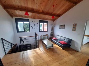 A modern living room with a wooden ceiling, a black leather sofa with red cushions, a white coffee table, and a staircase with a metal railing.