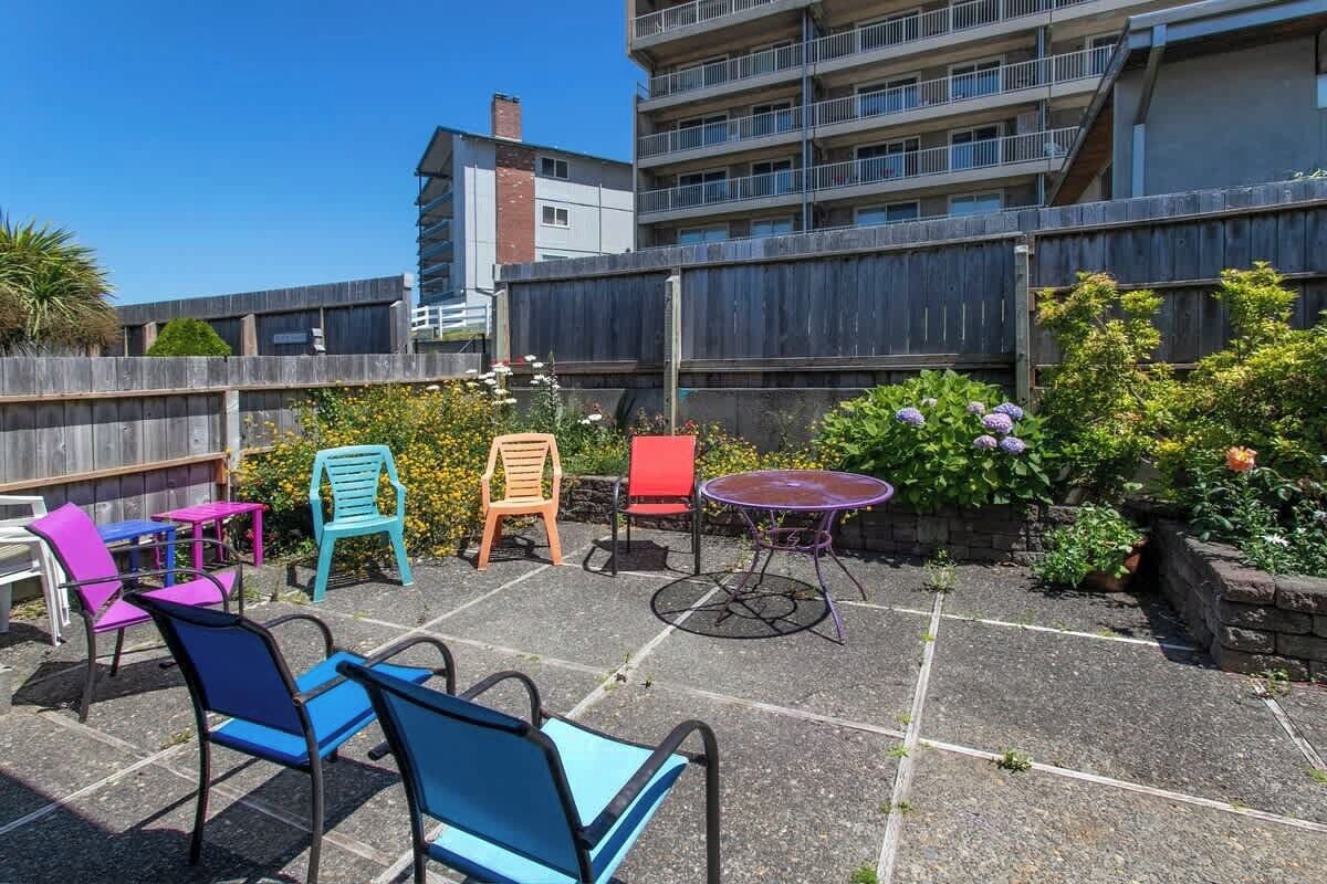 A rooftop patio with outdoor seating, including chairs and tables, surrounded by greenery and a view of a multi-story building.