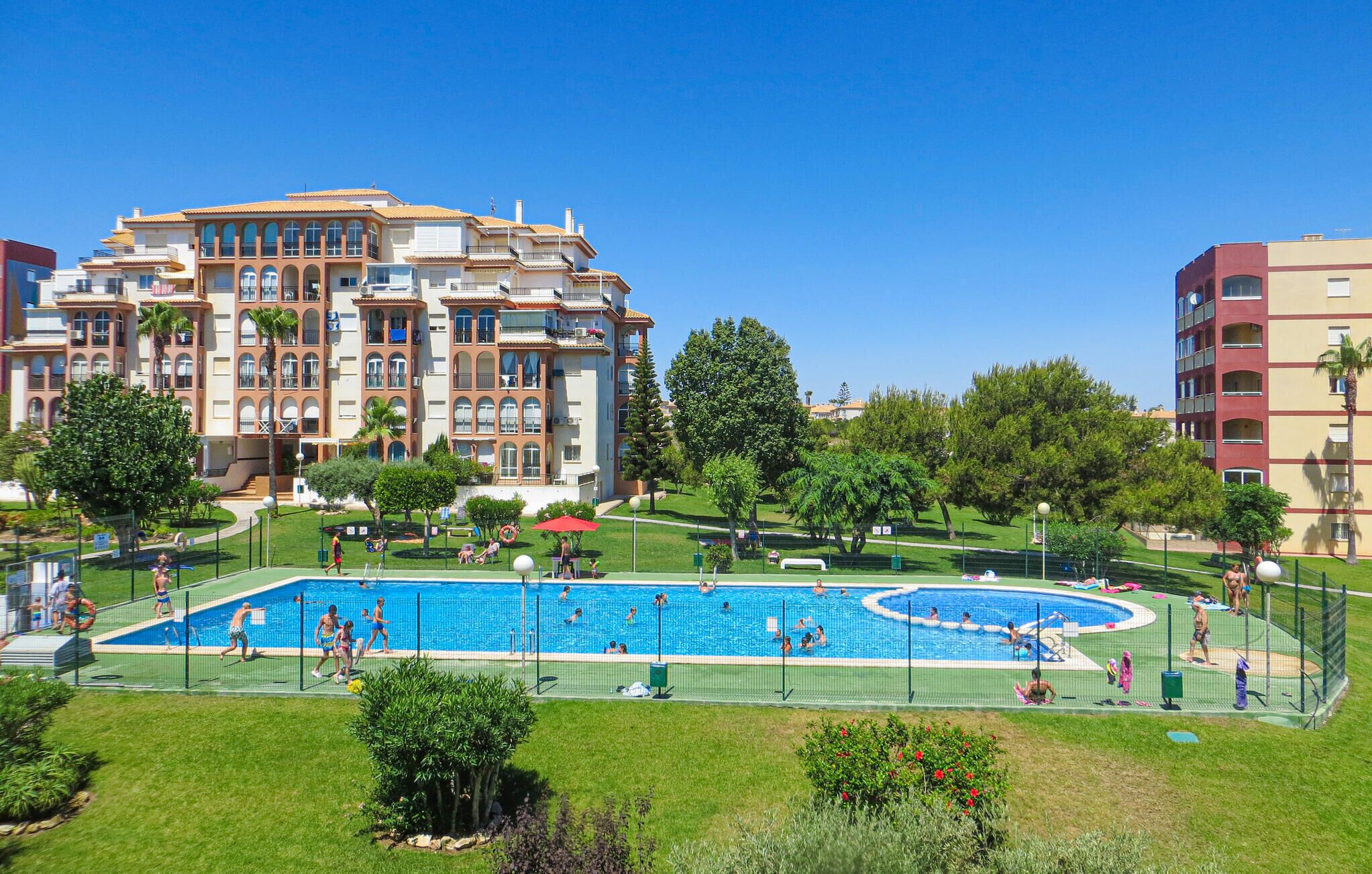 Piscina cercada por área delimitada, com pessoas nadando e brincando. Ao fundo, um edifício grande com várias varandas e um gramado bem cuidado.