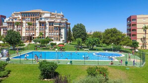 A swimming pool surrounded by a fenced area with people swimming and playing. In the background, there is a large building with multiple balconies and a well-maintained lawn. - Gorgeous apartment in Torrevieja (Torrevieja)