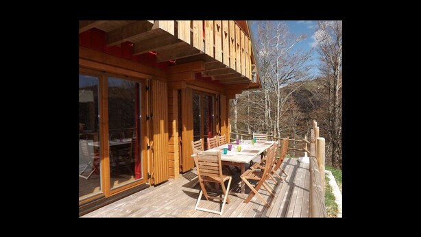 Une terrasse en bois avec une table et des chaises, de grandes portes vitrées et une vue sur les arbres et les collines.