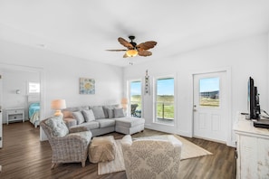 Un salon spacieux, doté d’un grand canapé d’angle, d’un ventilateur de plafond et offrant une vue sur l’extérieur grâce à une porte.