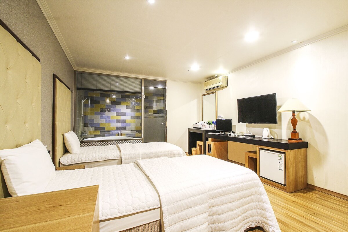 A hotel room with two beds, a desk with a TV, a mirror, and a window with a view of the city.