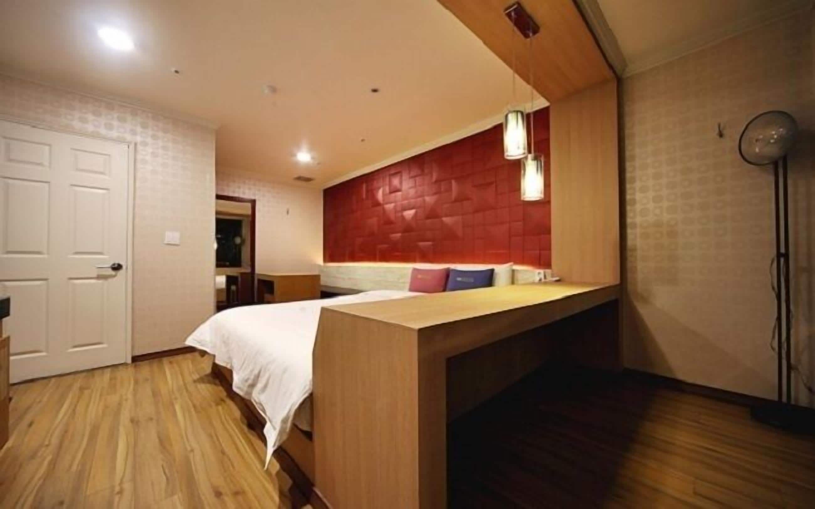 A bedroom with a bed, a wooden desk, a red headboard, and a floor lamp.