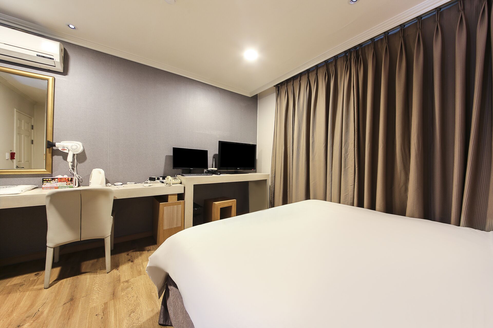 Habitación de hotel con cama, escritorio, computadora, dos monitores, secador de pelo y espejo.