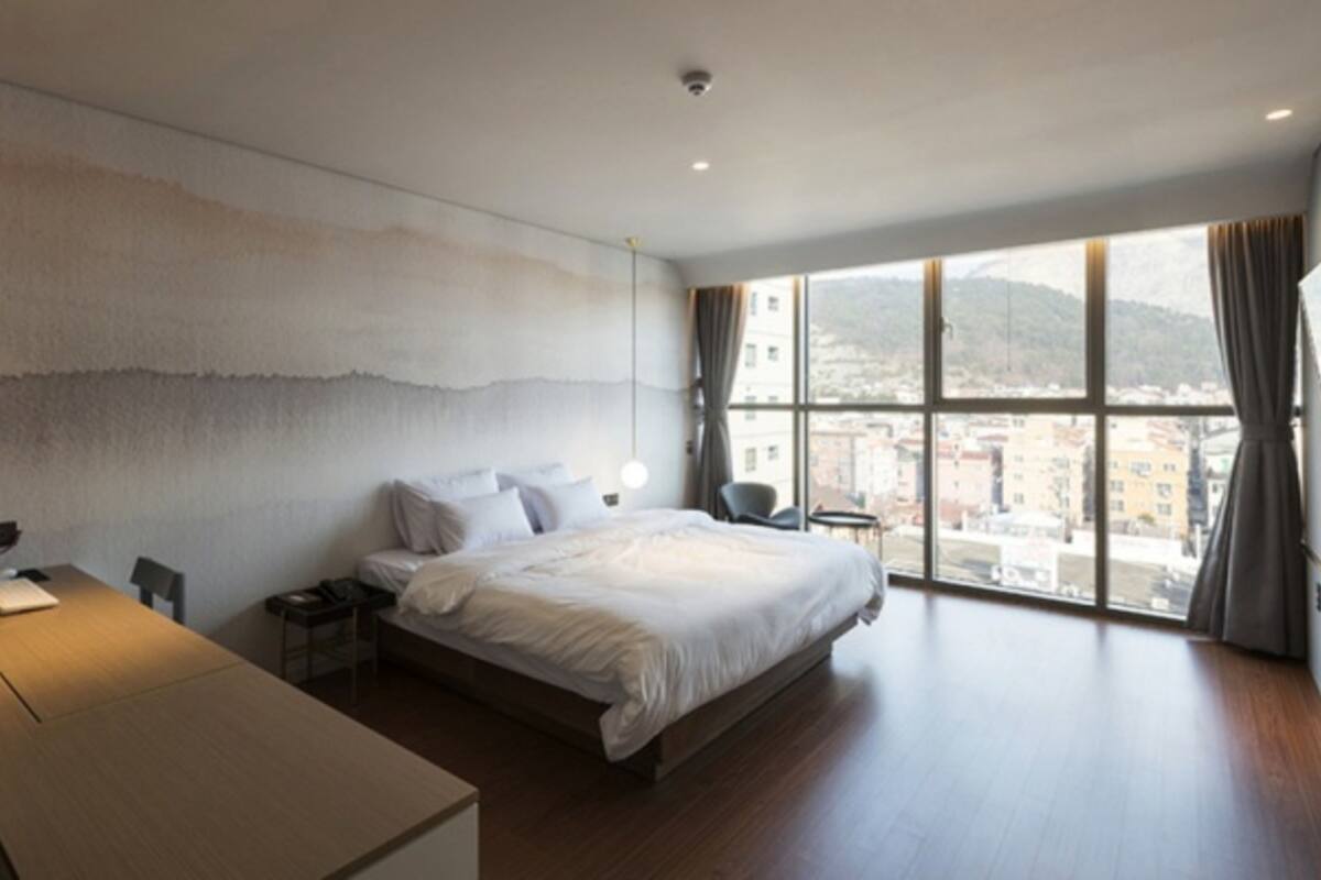 A modern hotel room with a large bed, a desk, and a view of the city through large windows.