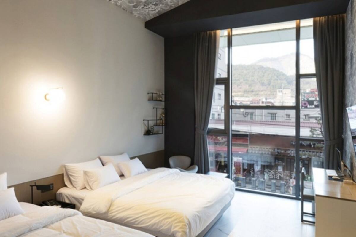 A modern hotel room with a large bed, a desk, and a view of the city through large windows.