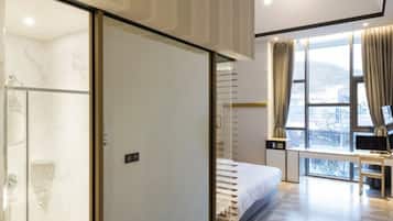 A modern hotel room with a large bed, a desk with a computer, and a view of the city through large windows.