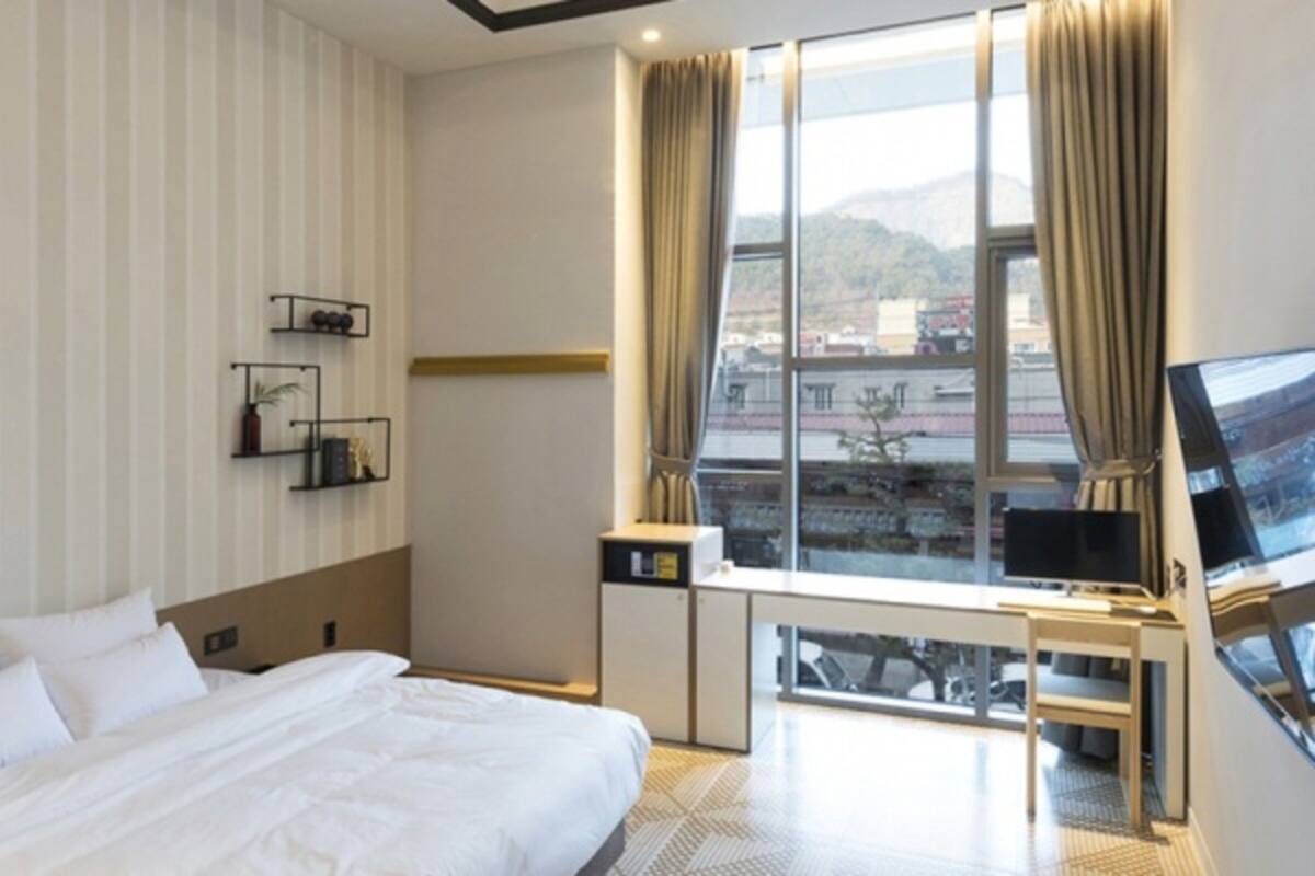 A hotel room with a large bed, a desk with a TV, a window with a view of the city, and a small shelf with decorative items.