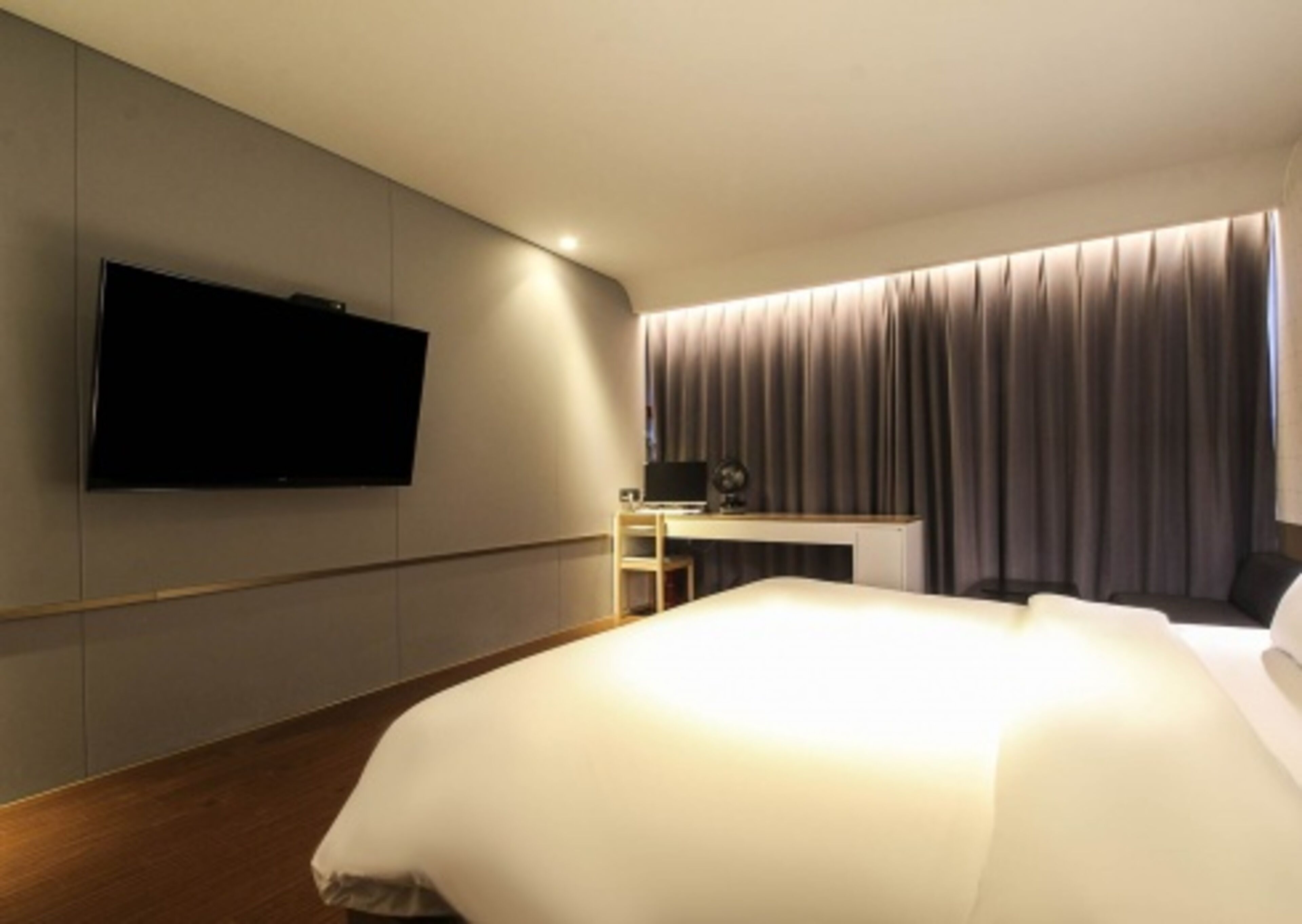 A modern hotel room with a large bed, a flat-screen TV mounted on the wall, a desk with a chair, and a large window with curtains.