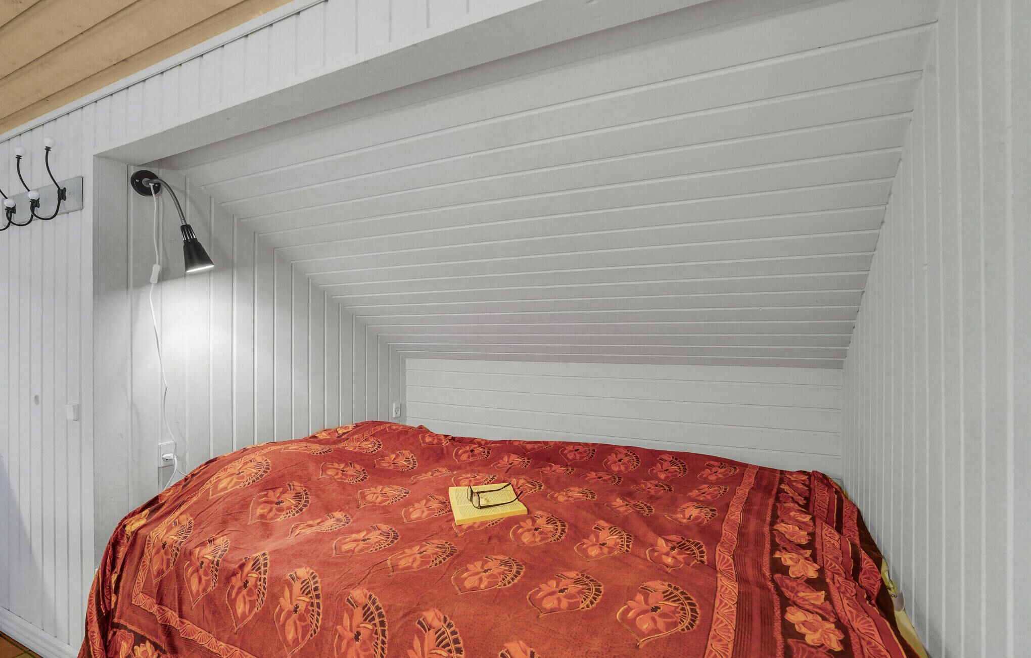 A small, narrow attic room with white wooden walls, a slanted ceiling, and a single bed with a red patterned bedspread.